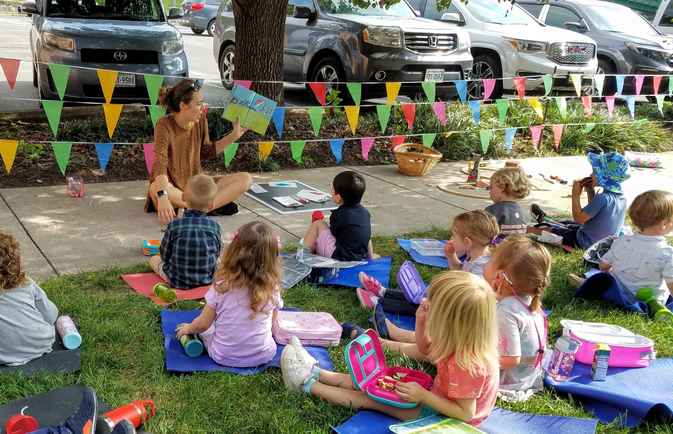 Children sit outside with an adults. Rainbow colored flags surround the area