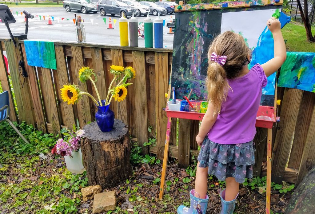 A child in a purple shirt paints on an easel. A vase with sunflowers is nearby