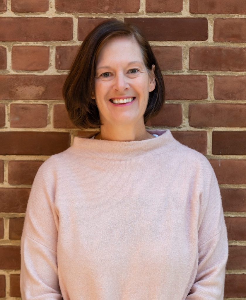 A smiling woman with chin length brown hair and wearing a pink top stands in front of a brick wall