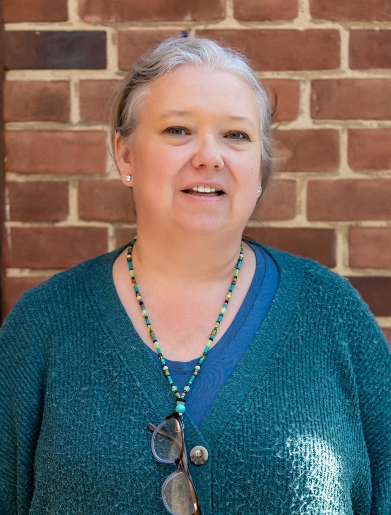 Headshot of a woman with gray hair pulled back standing in front of a brick wall