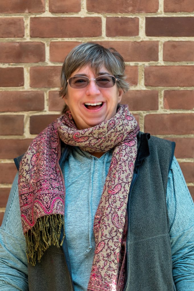 A smiling woman wearing a scarf stands in front of a brick wall