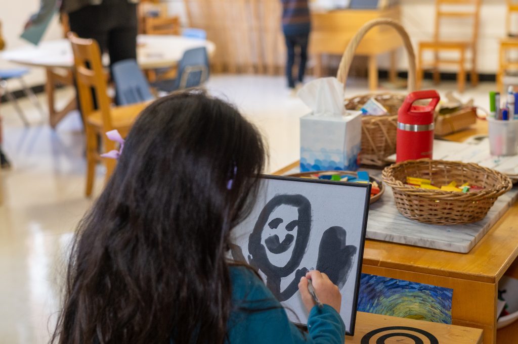 The back of a child's head. She is seated and painting a smilie face on a large piece of paper