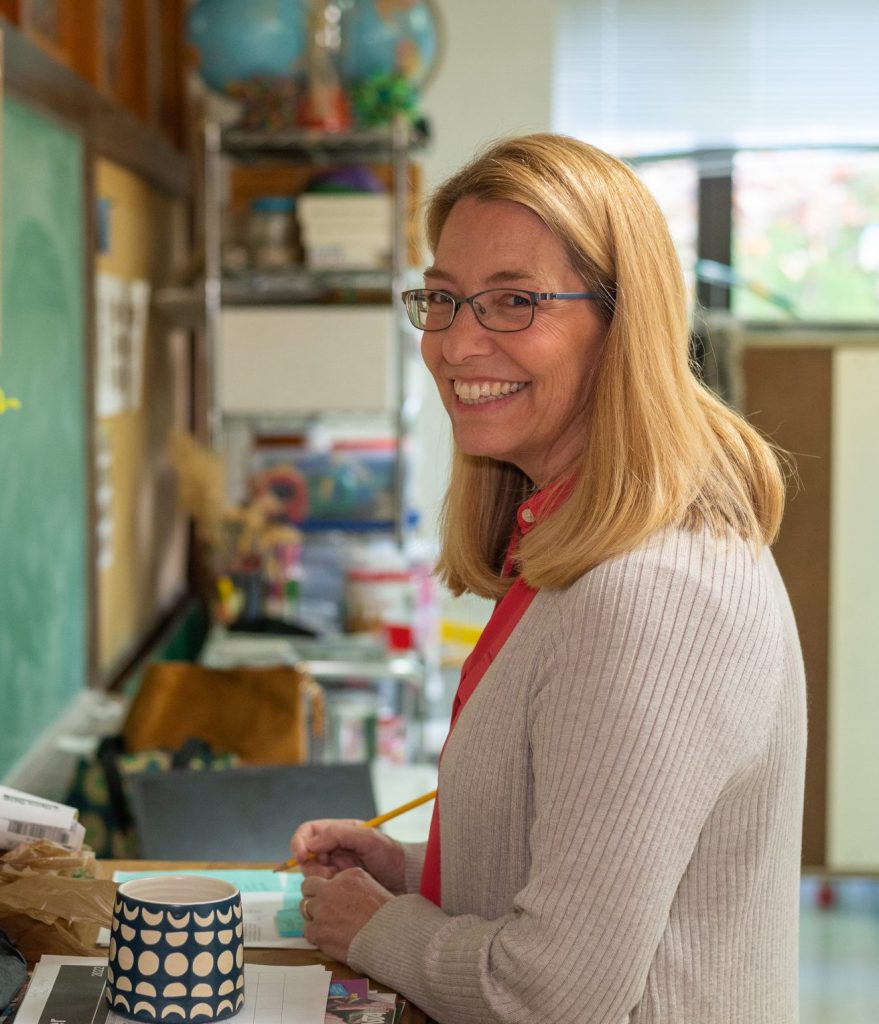 A smiling woman with shoulder length blonde hair wears a beige cardigan, holds a pencil