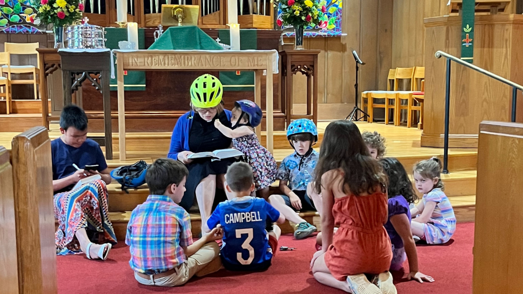 A pastor wearing a bicycle helmet sits on the floor and speaks with a group of children