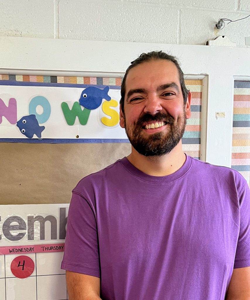 A smiling man with a beard stands in front of a bulletin board