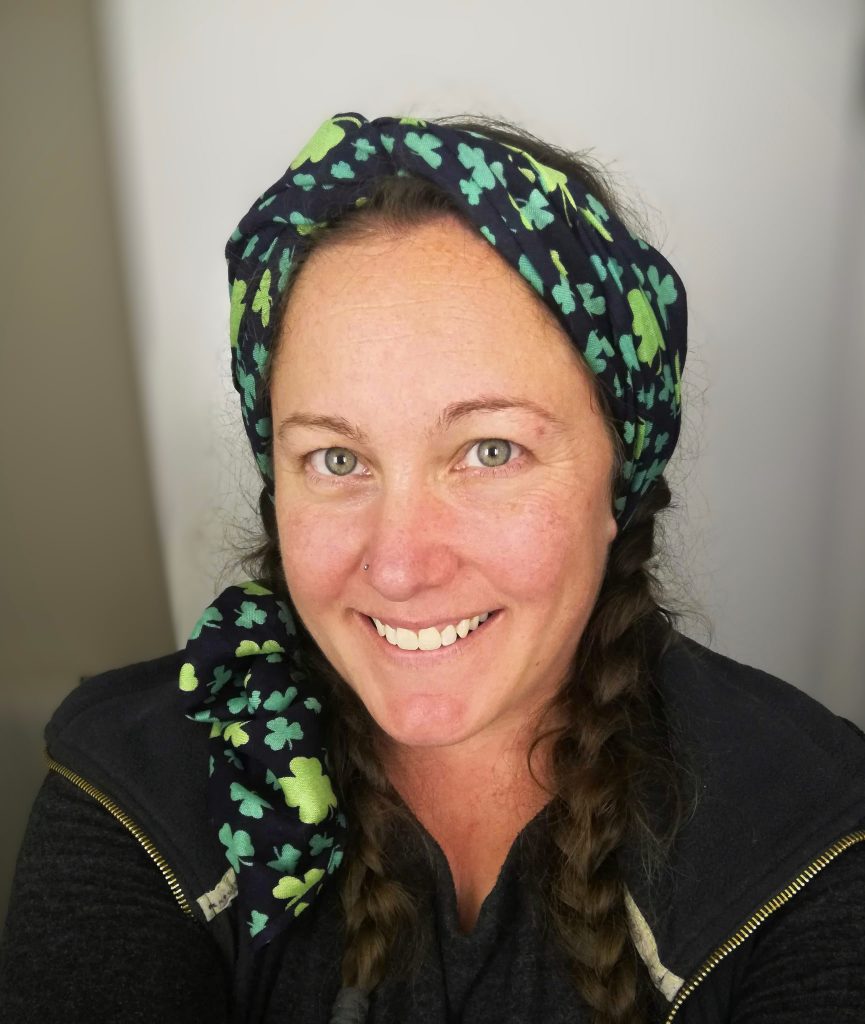 A smiling woman in a dark top and scarf covering her hair stands in front of a blank wall