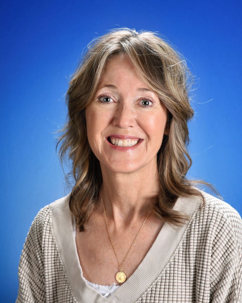 A smiling woman with shoulder length light brown hair in front of a blue background.