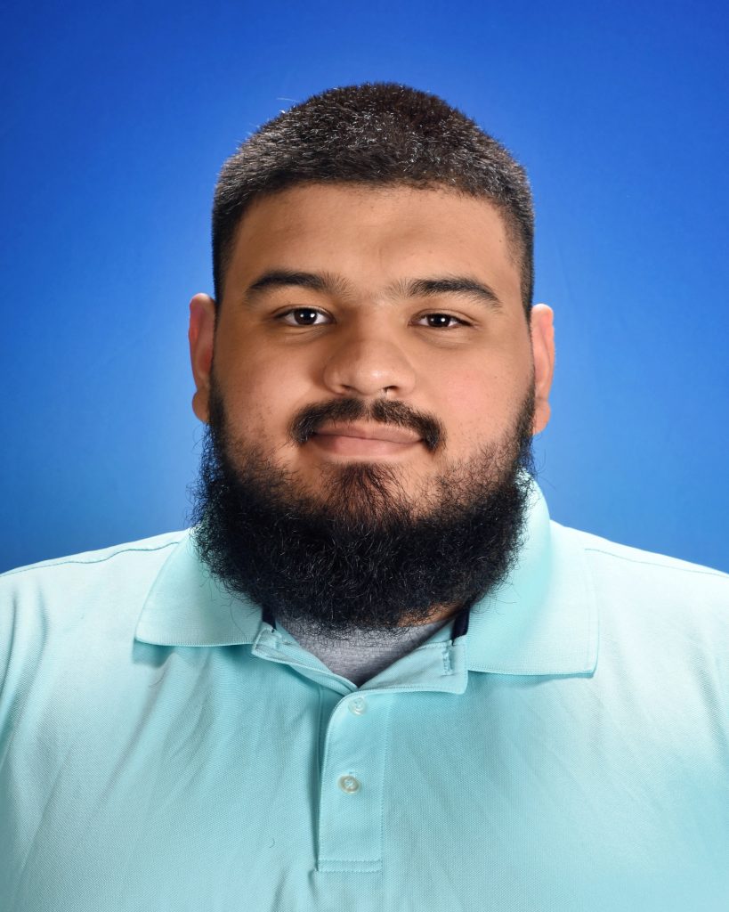 A man with a full beard and dark hair in front of a blue background