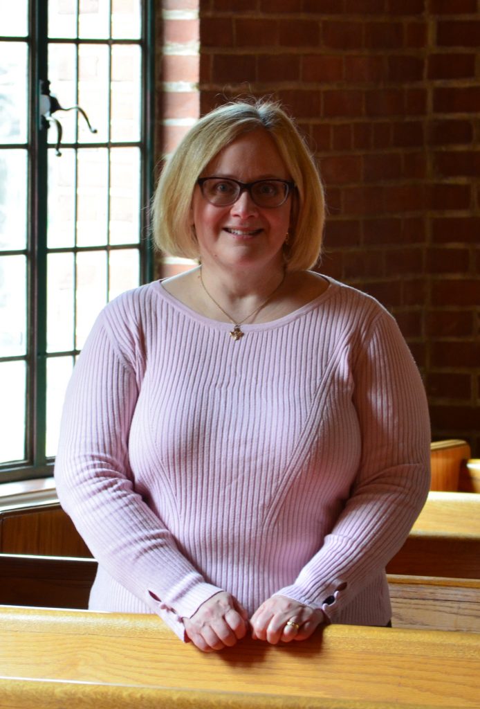 A smiling woman wearing a pink top stands indoors in front of a brick wall and a window