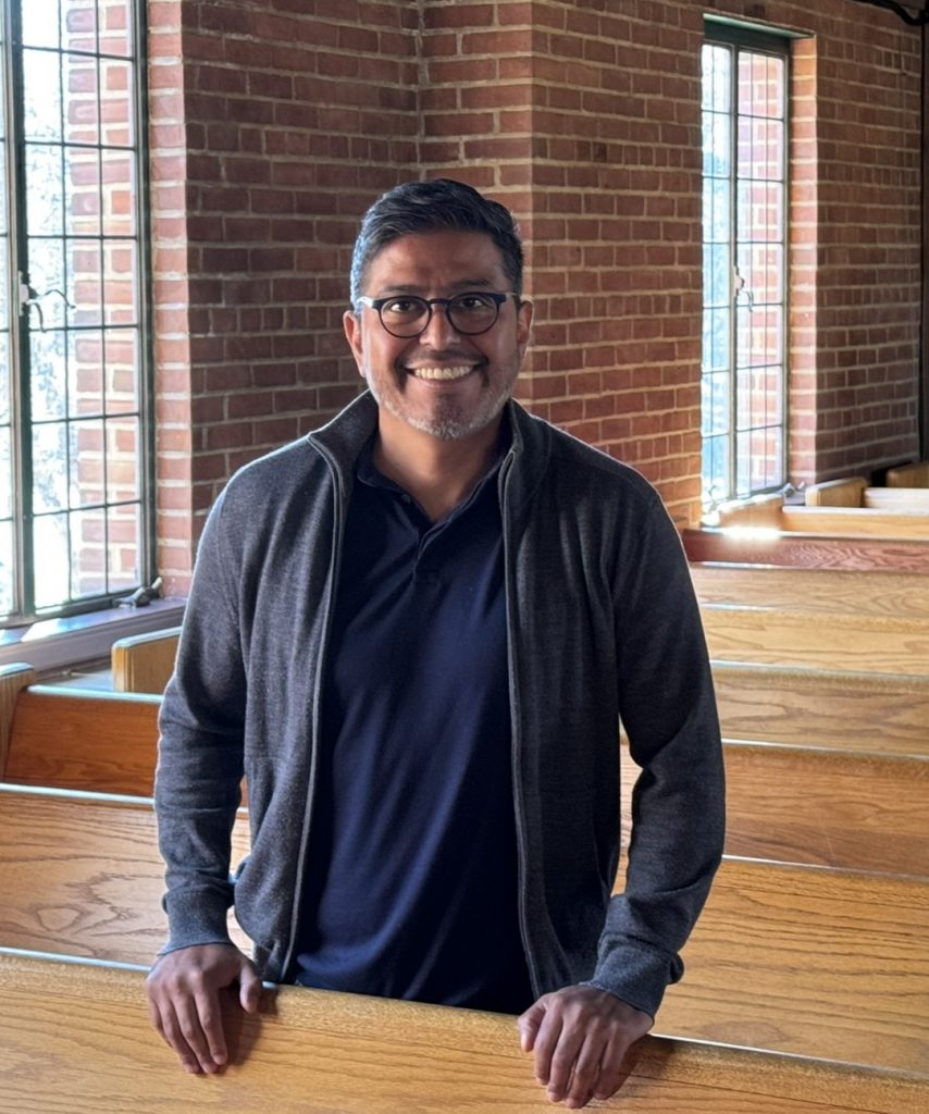 A smiling man wearing glasses stands indoors in front of a brick wall and two windows