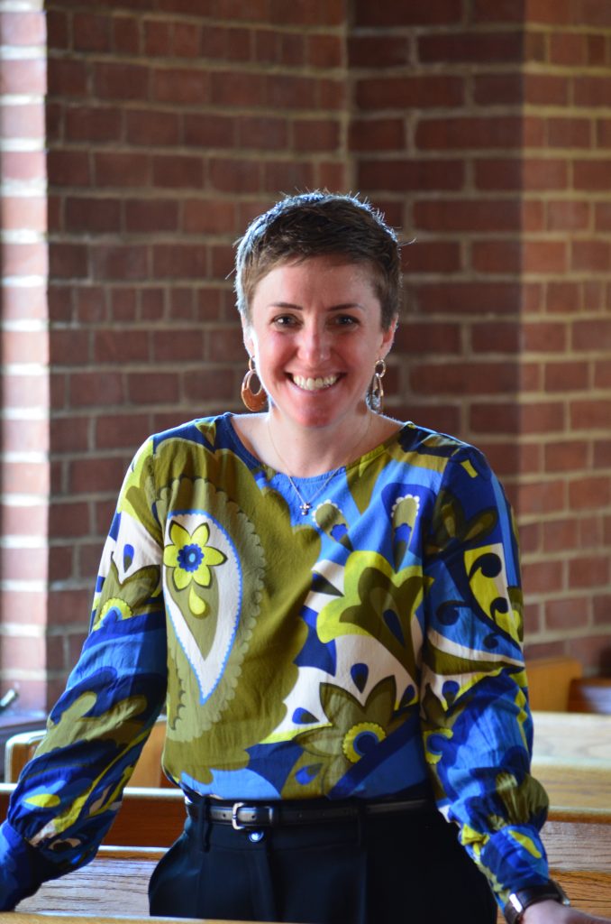 A smiling woman with short hair stands in front of a brick wall.