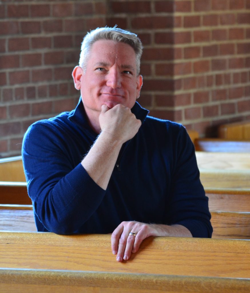 A smiling man with short hair leans his chin on his hand while seated in front of a brick wall