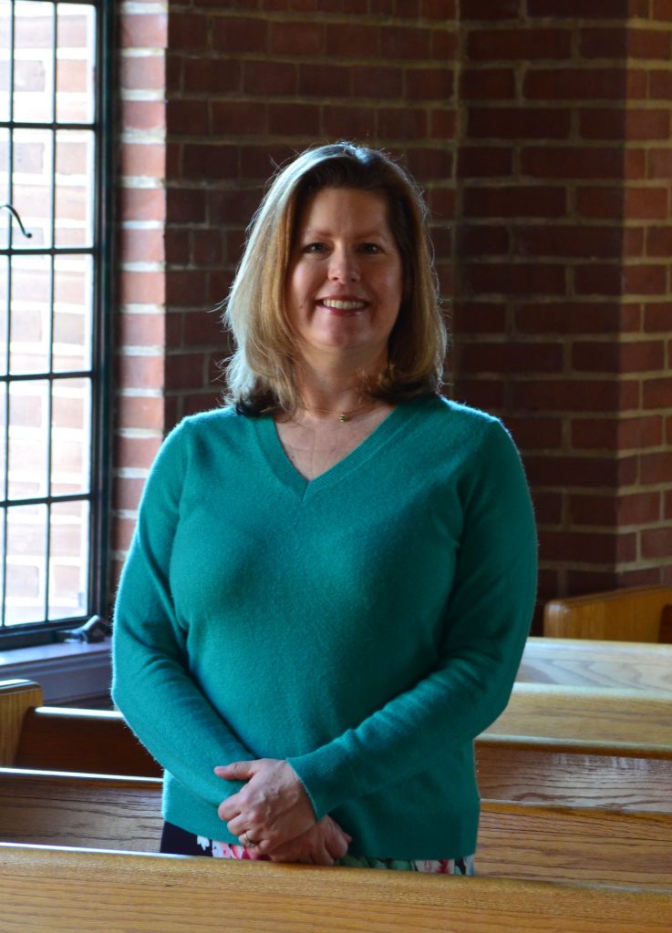 A smiling woman with shoulder length hair and a teal top stands in front of a brick wall