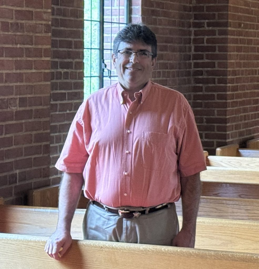 A smiling man in a pink shirt stands indoors in front of a brick wall and a window