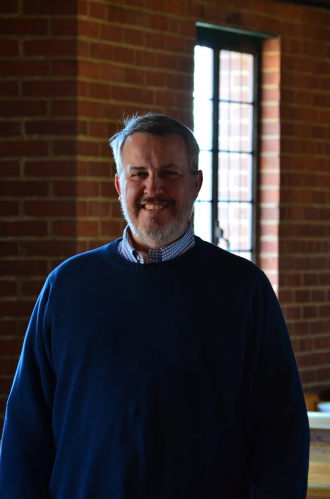 A man with short hair wearing a sweater stands in front of a window