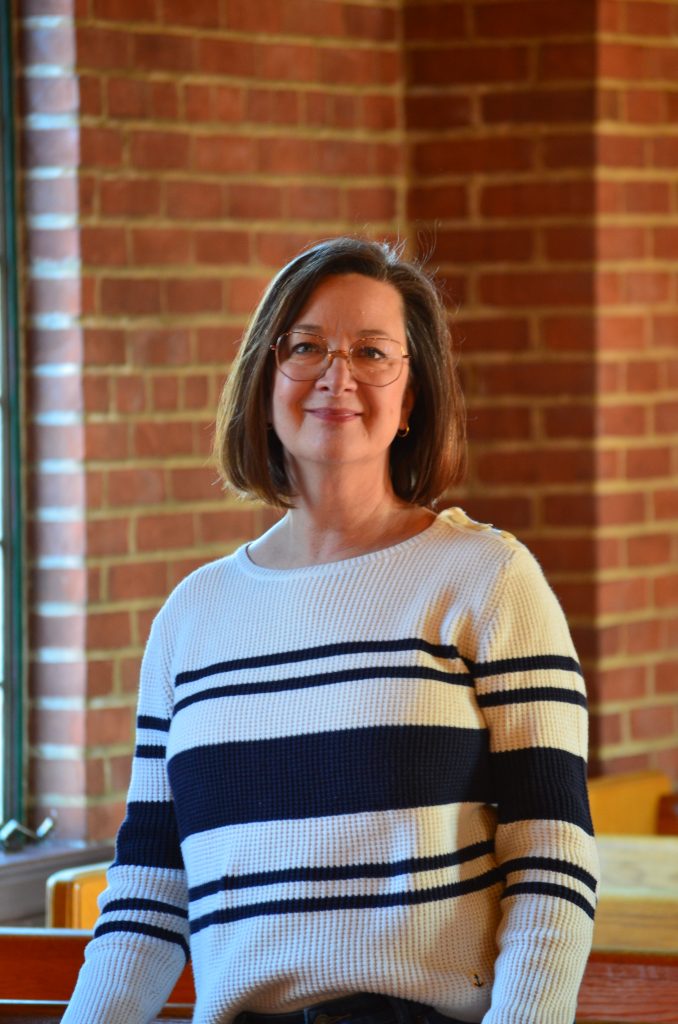 A smiling woman in a black and white striped top stands indoors in front of a brick wall