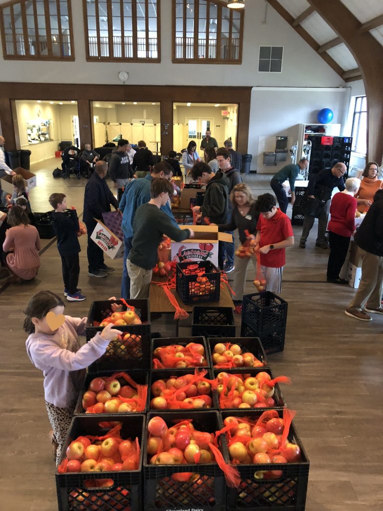A group of people stand around tables on which sit crates of apples