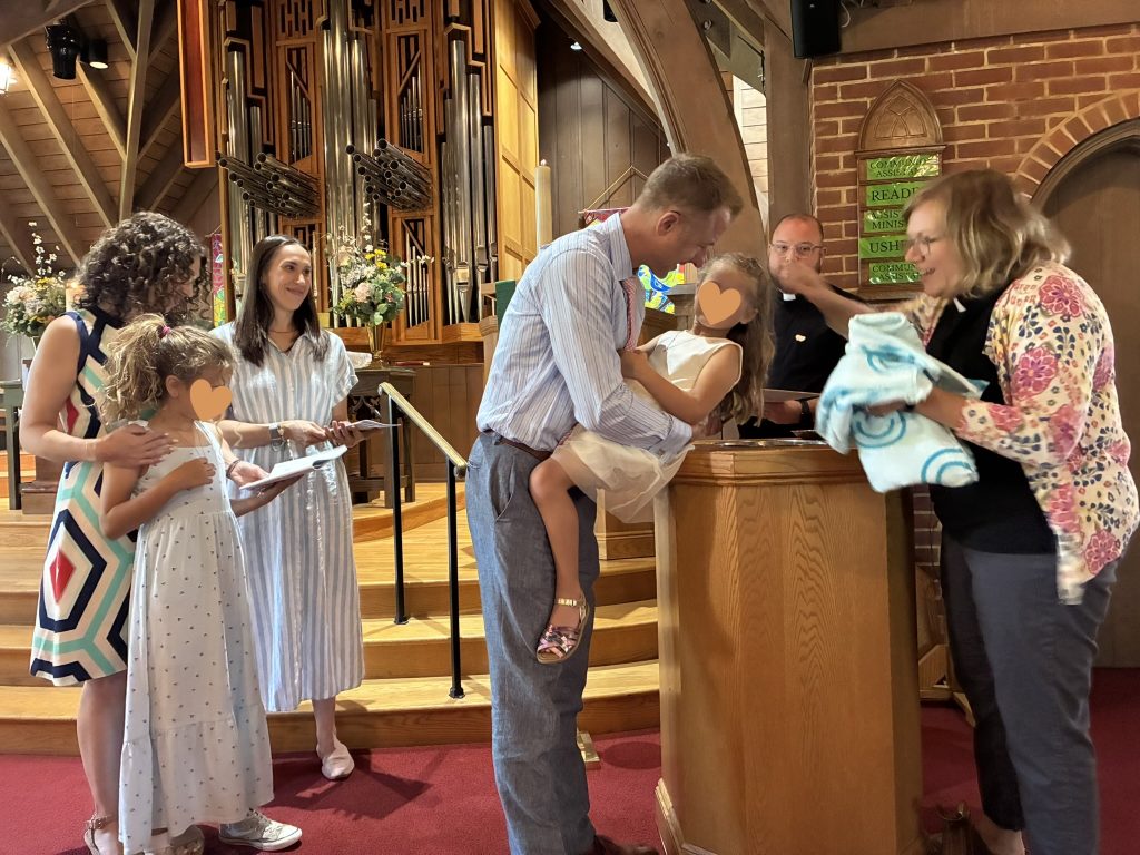 A man holds a young girl over a baptismal font while the pastor reaches out a hand