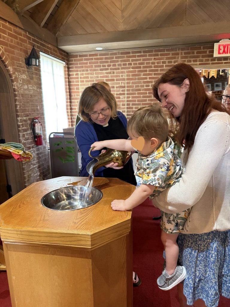 Two women hold a small child over a baptismal font; the child reaches for the water